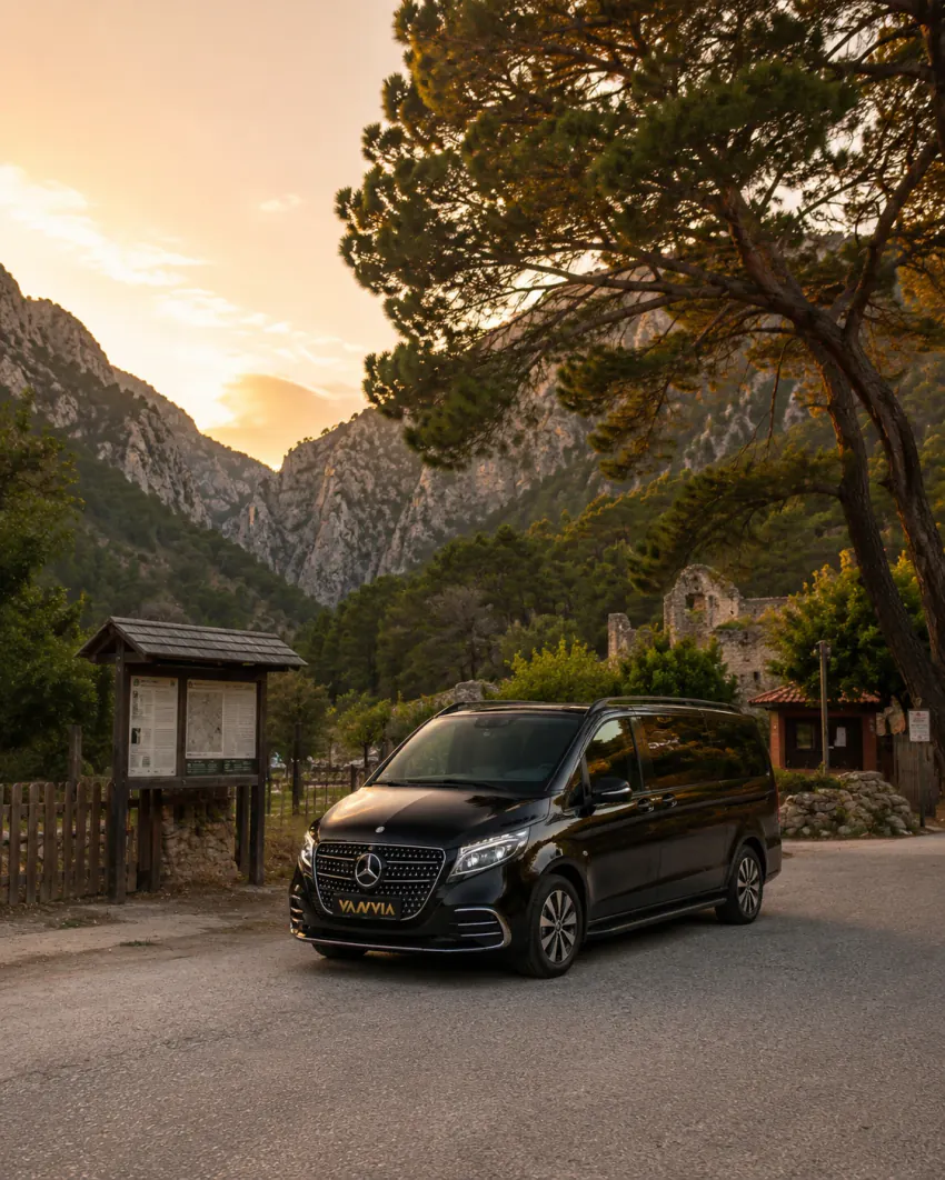 Olimpos Antik Kenti'nin genel giriş ve otopark alanında, bilgilendirme panosu ve arkada antik kalıntıların göründüğü bir noktada gün batımında duran siyah Vanvia VIP transfer aracının ön görünümü