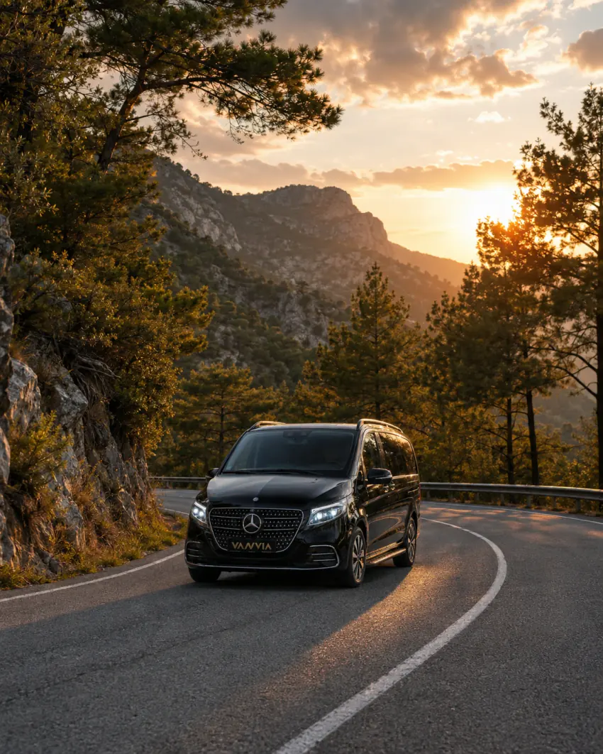 Olimpos çevresindeki çam ormanı içinde kavisli bir dağ yolunda, korkuluk ve ağaçların arasından süzülen gün batımı ışığıyla ilerleyen siyah Vanvia VIP transfer aracının ön görünümü