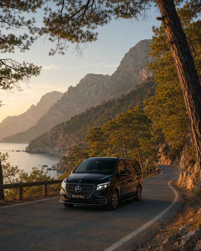 Olimpos yakınlarında ormanlık bir yamaç yolunda, ağaçların arasından aşağıda bir koyun göründüğü bir noktada altın ışıkta ilerleyen siyah Vanvia VIP transfer aracının ön görünümü