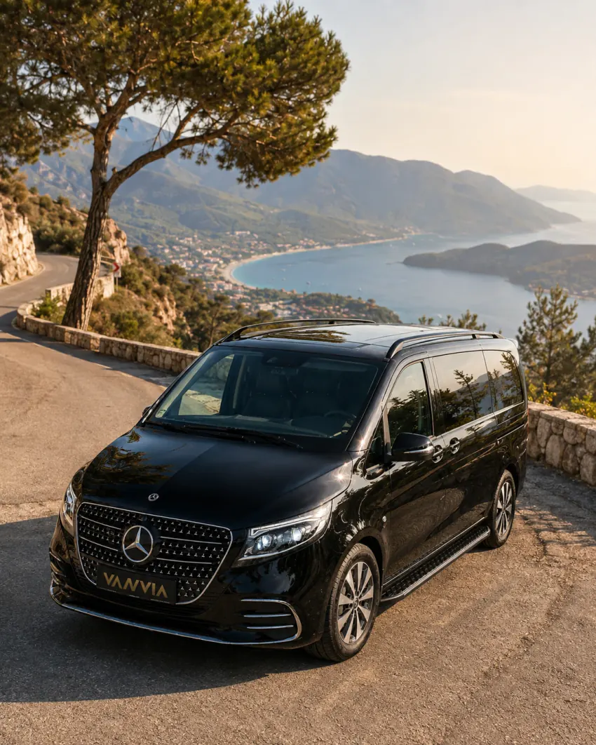 A front three-quarter view of a black Vanvia VIP transfer vehicle driving a mountain road near Ölüdeniz in golden daylight, with a bay view below