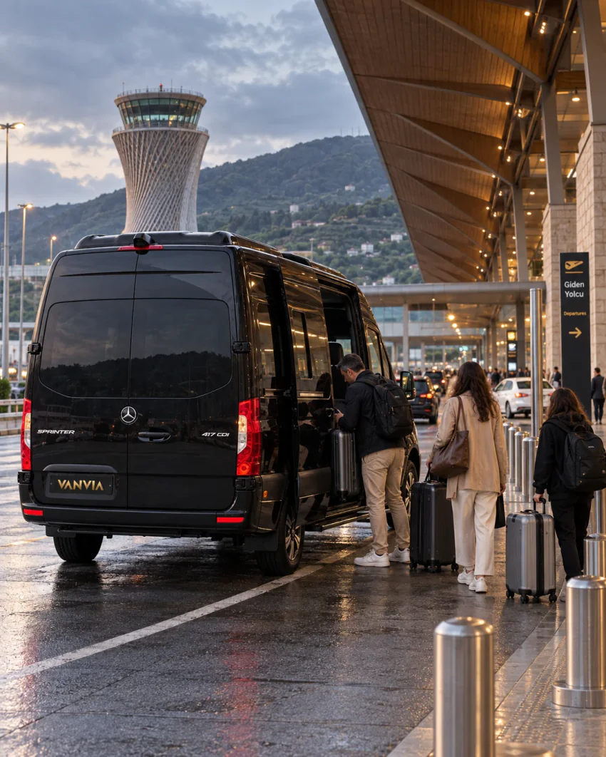 حافلة صغيرة سوداء لخدمة Vanvia VIP transfer تنتظر وبابها الجانبي مفتوح تحت مظلة صالة مطار رايزة-أرتفين بينما يصعد الركاب ومعهم حقائبهم ذات العجلات، ويظهر برج المراقبة في الخلفية