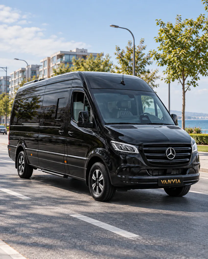 Ein schwarzes Vanvia-VIP-Transferfahrzeug auf einer breiten, von Bäumen und Wohnhäusern gesäumten Stadtstraße, am Ende ist das Meer zu sehen