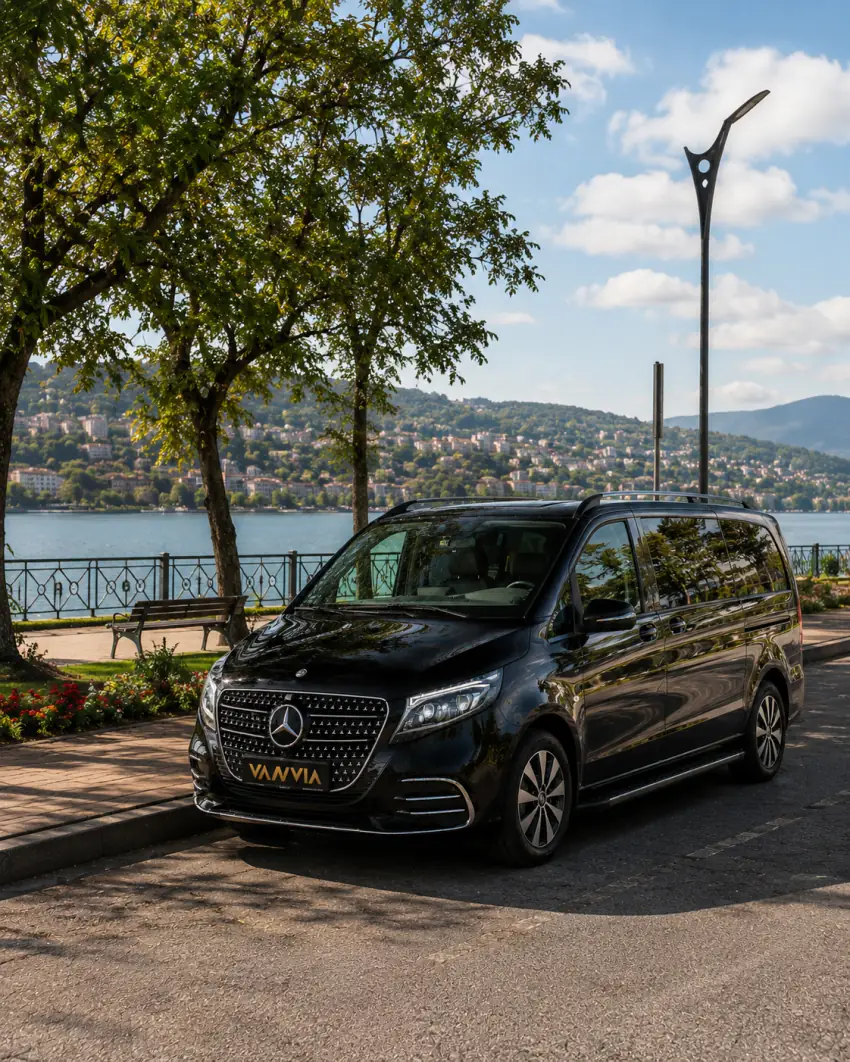 Göl kenarındaki taş döşeli, korkuluklu bir yürüyüş yolunda duran siyah Vanvia VIP transfer aracının ön çapraz görünümü, karşı kıyıda tepeye yayılmış bir yerleşim