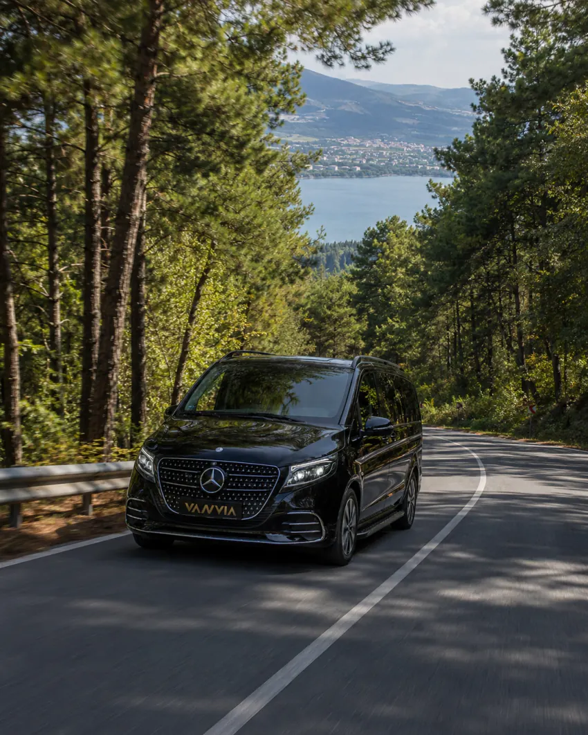 Ağaçlarla çevrili, aşağı inen bir yolda ilerleyen, ilerisinde ağaçların arasından bir gölün göründüğü siyah Vanvia VIP transfer aracının ön çapraz görünümü
