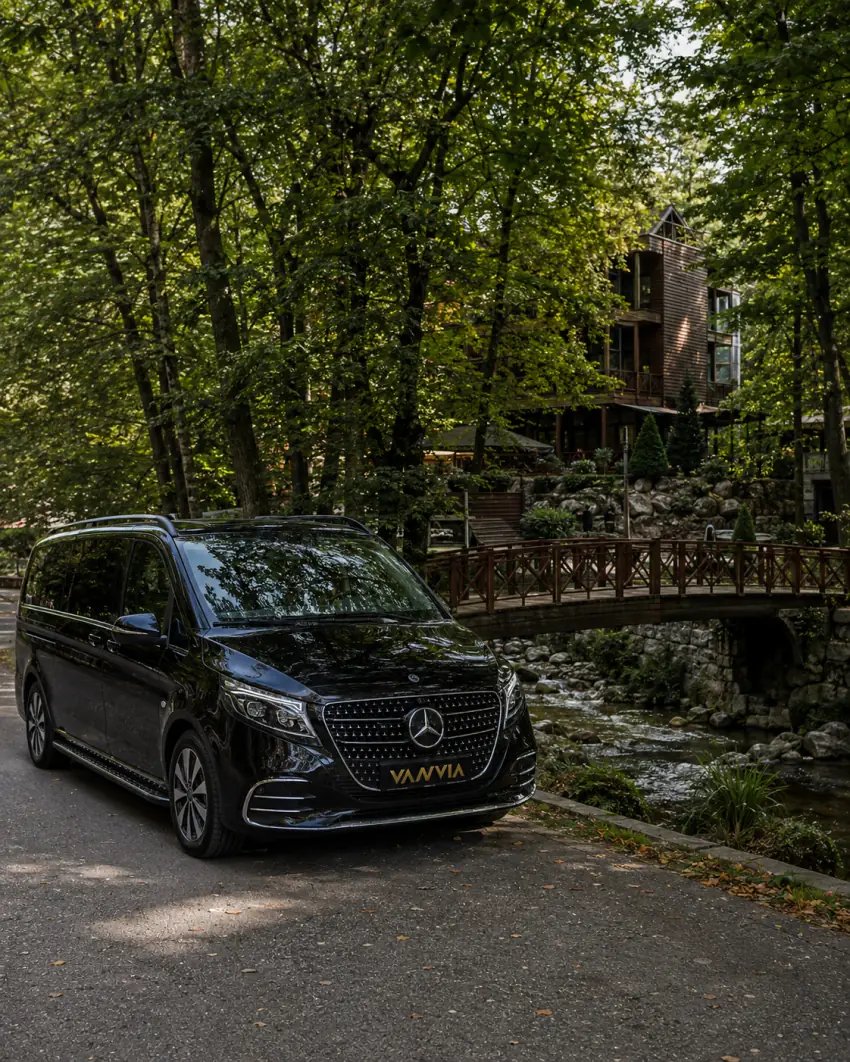 Bir derenin üzerindeki ahşap köprünün yakınında duran siyah Vanvia VIP transfer aracının görünümü, ağaçların arasında ahşap cepheli bir bina eşliğinde