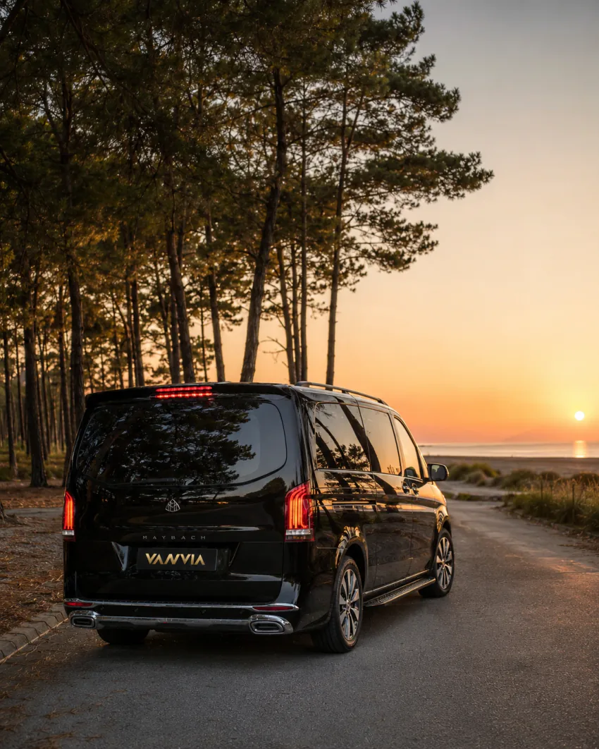 Sarıgerme'de gün batımında çam ağaçlarıyla çevrili asfalt plaj erişim yolunda duran siyah Vanvia VIP transfer aracının arka görünümü, ileride sahil ve deniz manzarası