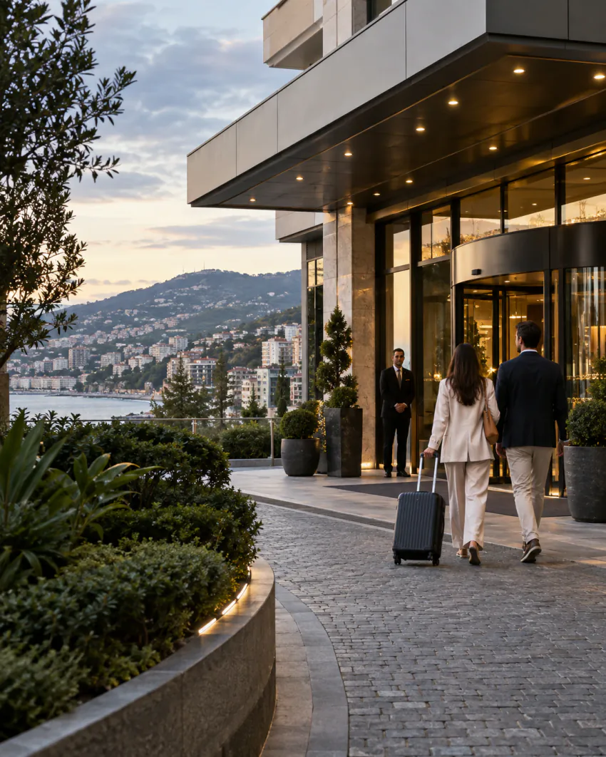Kendi valizini çeken bir çift, modern bir otel girişine doğru yürüyor; girişte bir görevli bekliyor, arka planda tepeye yayılan kıyı şehri manzarası görülüyor; otel adı veya markası görünmüyor, araç yok