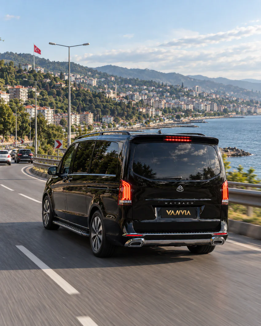 Vanvia VIP transfer aracı, deniz manzaralı kıvrımlı bir kıyı yolunda ilerliyor; arka planda tepeye yayılan şehir dokusu ve bir Türk bayrağı görülüyor
