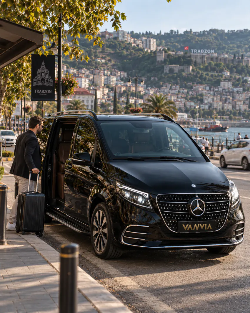 Siyah Vanvia VIP transfer aracı Trabzon'da bir kıyı caddesinde duruyor; bir yolcu açık kapıdan valizini alıyor, arka planda tarihi taş dokulu bir yapı ve şehrin adını taşıyan bir tabela görülüyor