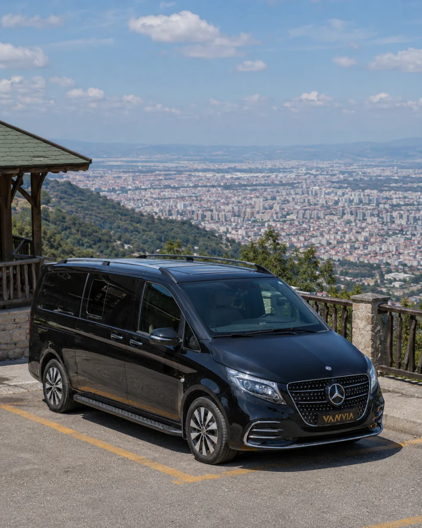 Aşağıda geniş bir şehir manzarasının yayıldığı, korkuluklu bir seyir noktasında duran siyah Vanvia VIP transfer aracının ön çapraz görünümü