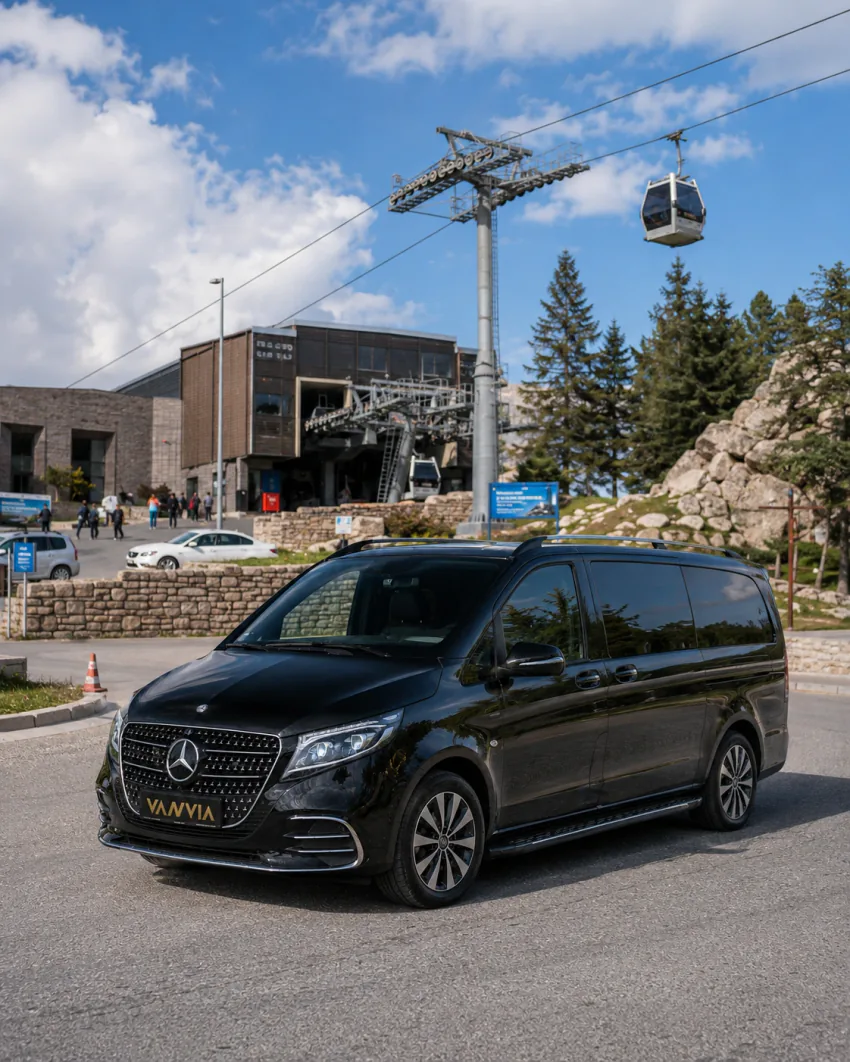 Bir teleferik hattının alt istasyon binası ve üzerinden geçen bir kabinin göründüğü açık otoparkta duran siyah Vanvia VIP transfer aracının ön görünümü