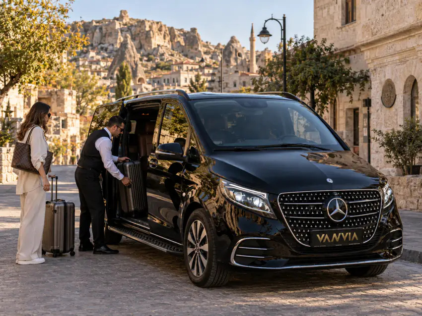 Arka kapısı açık siyah Vanvia VIP transfer aracının bagajına bir valizi yerleştiren şoför, yanında bagajlarıyla bekleyen bir kadın ve bir erkek, arka planda Ürgüp'ün eski kasaba silüeti ve bir sokak lambası
