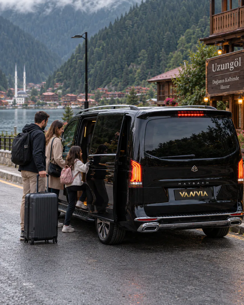 Bir aile, kendi valiziyle Vanvia VIP transfer aracının açık kapısından binmeye hazırlanıyor; arka planda 'Uzungöl' yazılı bir tabela, ahşap köy evleri, göl ve cami görülüyor