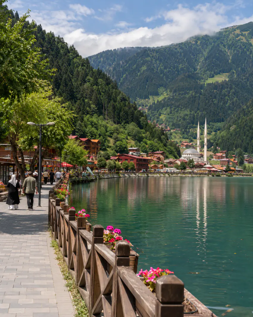 Uzungöl'de gölün kenarındaki ahşap korkuluklu yürüyüş yolunda insanlar yürüyor; arka planda iki minareli bir cami, ahşap köy evleri ve ormanlık dağlar görülüyor; araç yok