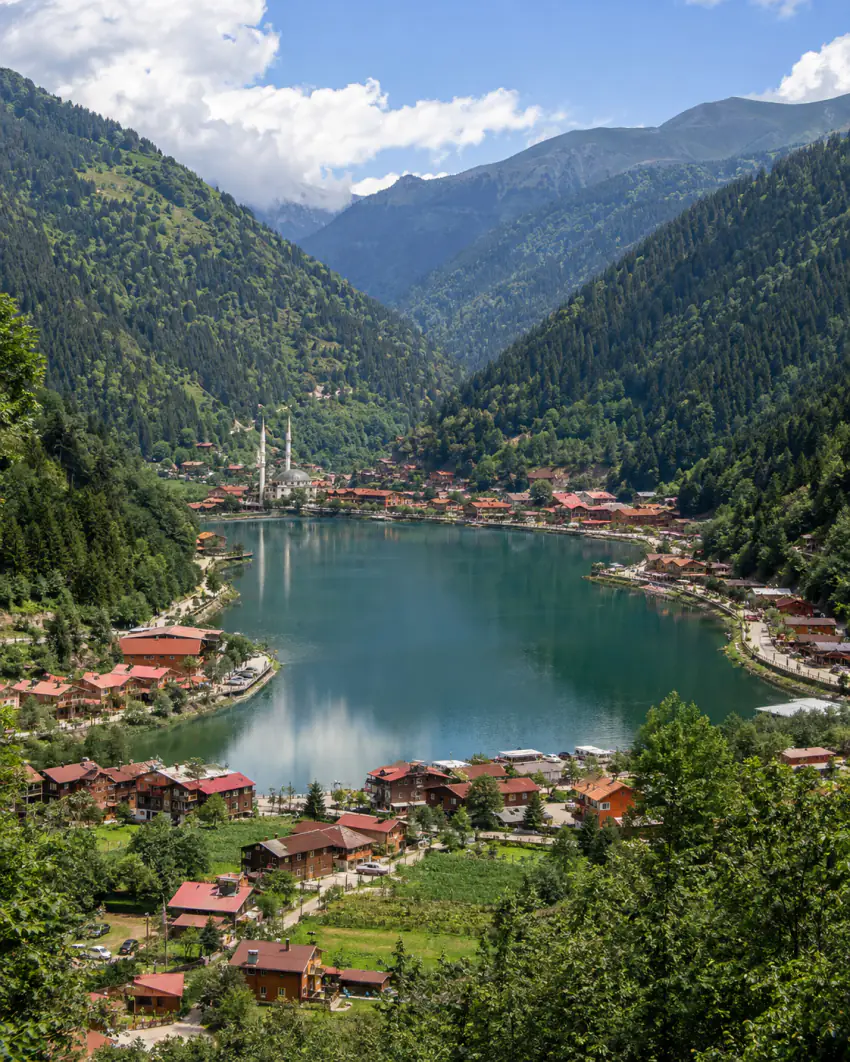 Uzungöl'ün yükseklerden çekilmiş panoramik görünümü; zümrüt yeşili göl, dik ormanlık vadi, kırmızı-kahverengi çatılı köy/konaklama kuşağı ve iki minareli bir cami görülüyor; araç yok