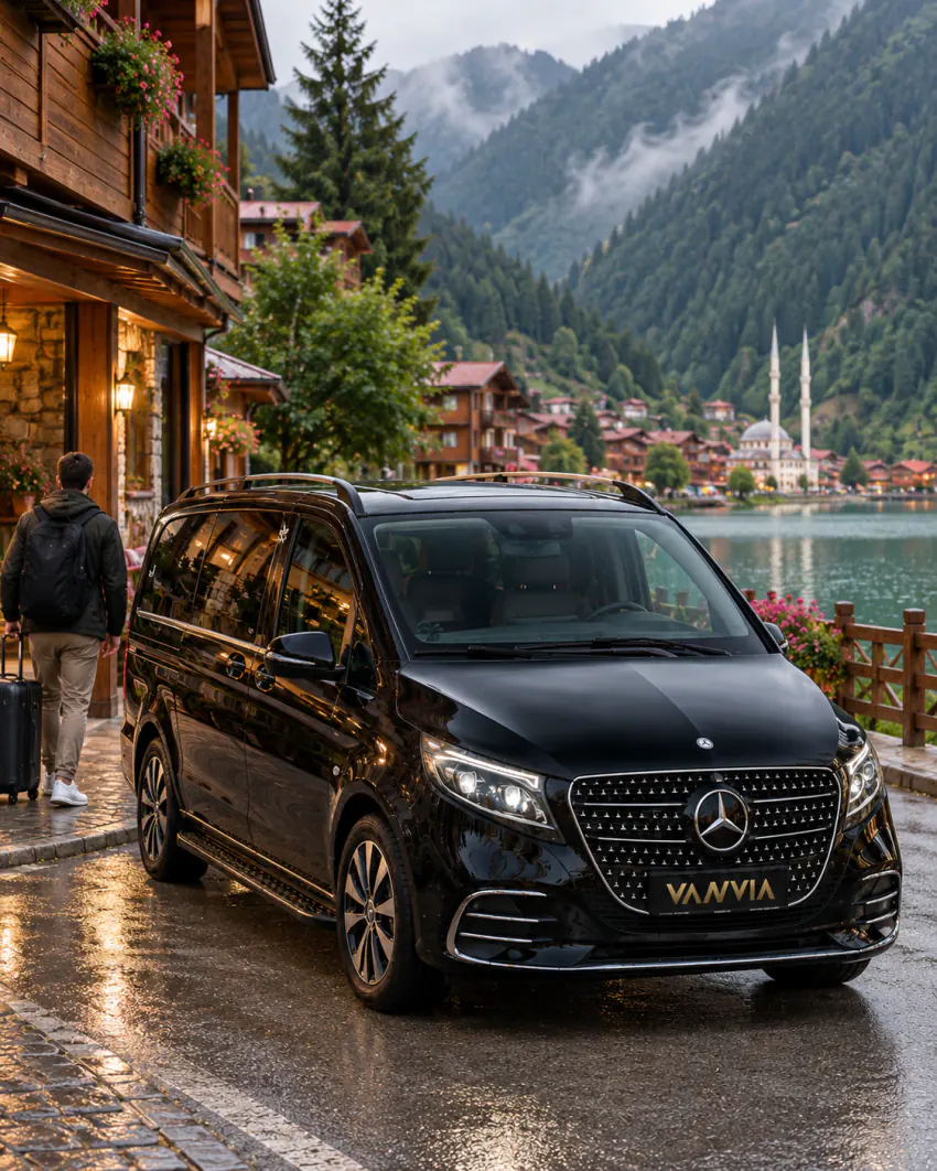 Vanvia VIP transfer aracı, Uzungöl'ün konaklama bölgesinde ahşap cepheli bir binanın önünde duruyor; bir yolcu kendi valiziyle binaya doğru yürüyor, arka planda göl, cami ve köy manzarası görülüyor