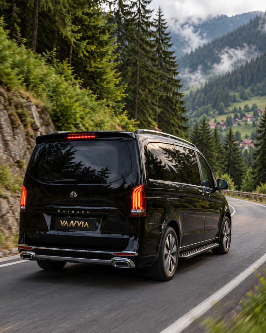 Vanvia VIP transfer aracı, ormanlık bir dağ yamacındaki kıvrımlı asfalt bir yayla yolunda ilerliyor; arka planda uzakta dağ evleri ve yeşil vadi görülüyor