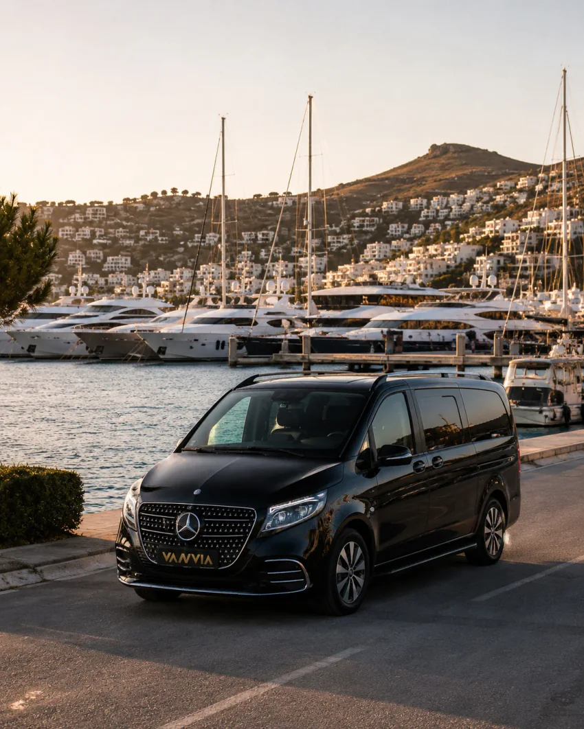 A black Vanvia VIP transfer vehicle waiting in front of the yachts at Yalıkavak Marina, with white hillside houses behind